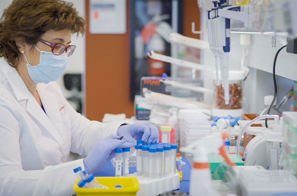 A woman wearing a lab coat, mask, and gloves, works at a lab bench doing mRNA research